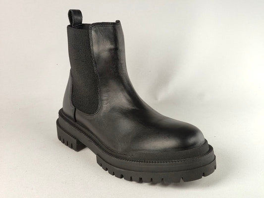 A black leather Chelsea boot with a chunky lug sole and elastic side gore, photographed on a light gray studio background.