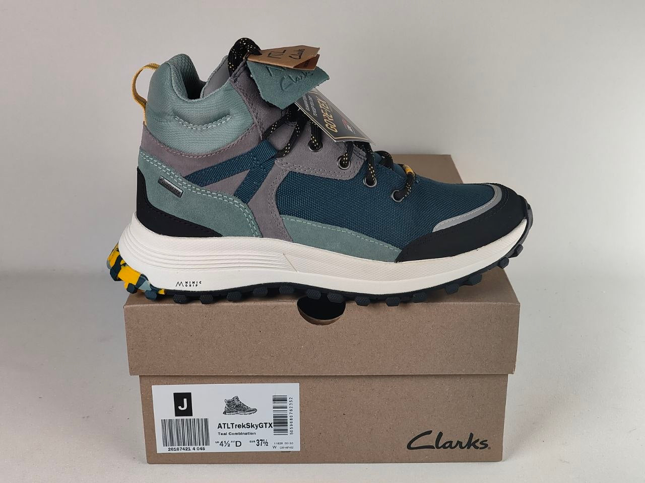 A Clarks outdoor hiking shoe in teal, gray, and black, displayed on top of its brown cardboard shoebox against a neutral light gray background.