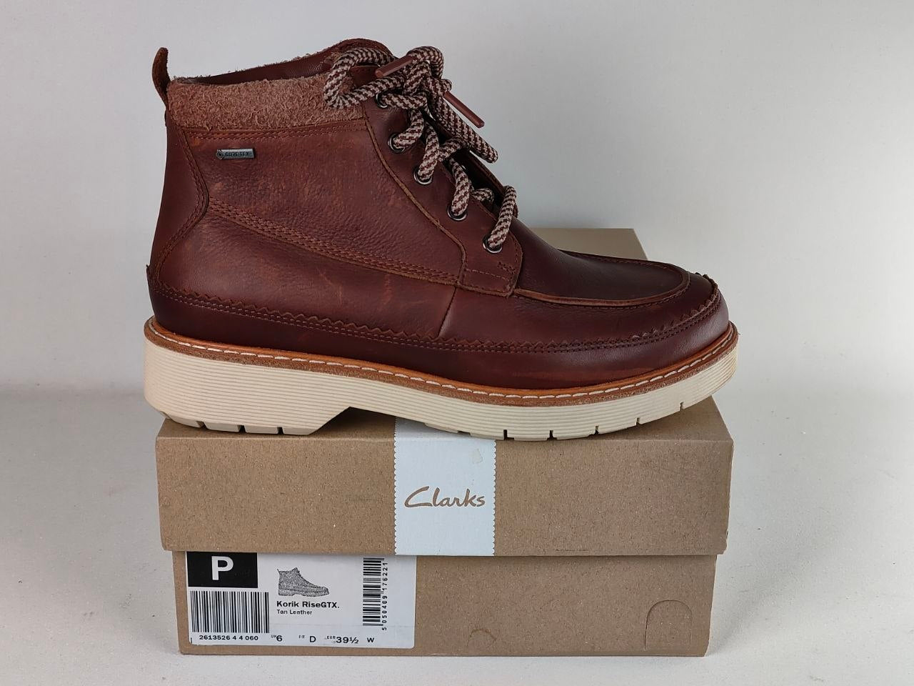 A brown leather ankle boot with patterned laces, resting on top of its brown Clarks shoebox against a light gray background.