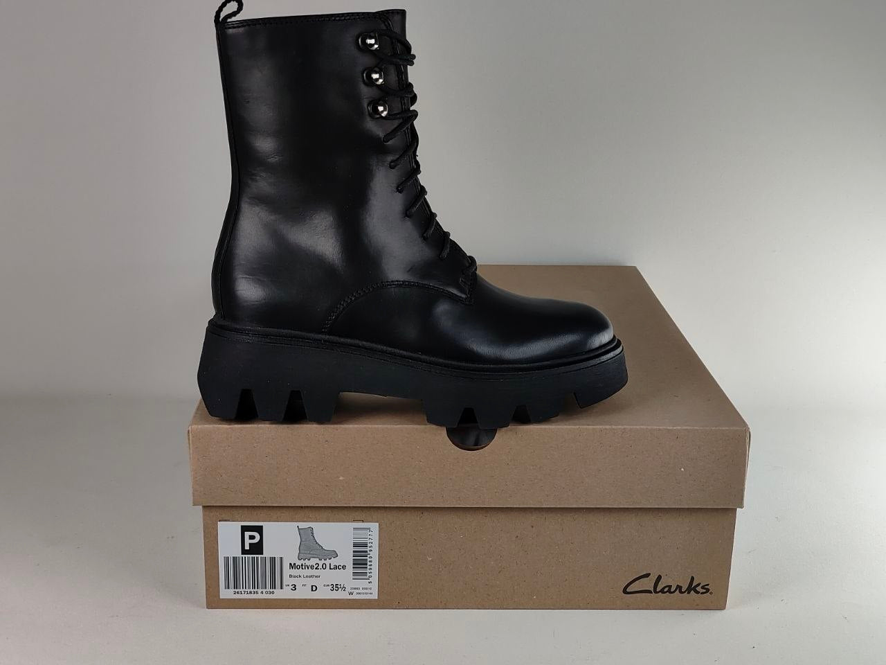A black leather lace-up ankle boot with a chunky lug sole, displayed on top of a brown Clarks shoebox against a neutral gray background.