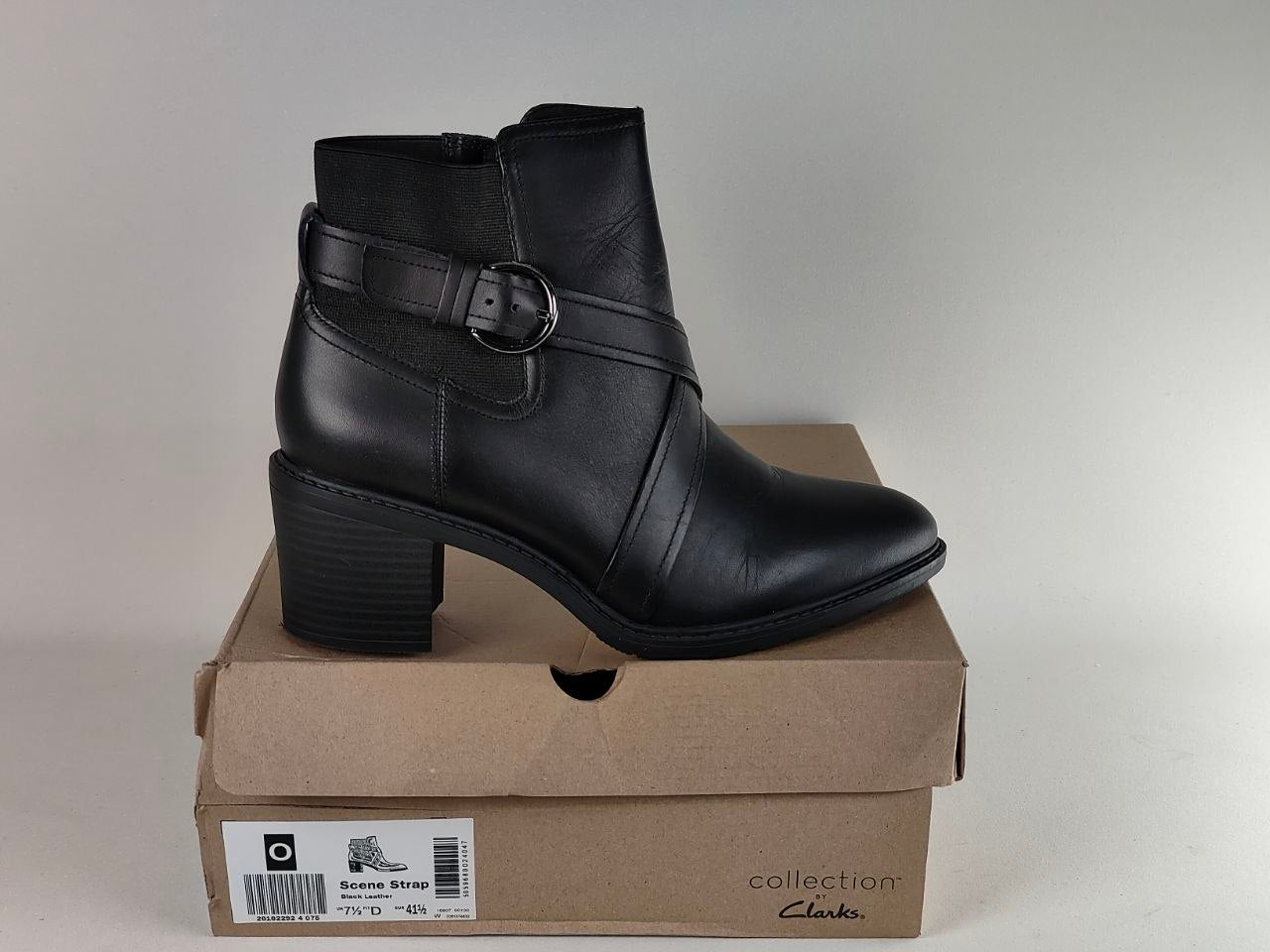 A black leather ankle boot with a side buckle strap, displayed on top of its brown cardboard shoebox. The box features the 'collection Clark's' logo and a size label for the 'Buckled Strap' model.
