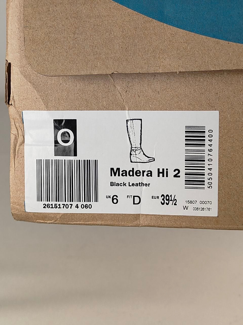 A close-up of a brown cardboard shoe box showing a white product label for a Madera Hi 2 boot in black leather, with a size 6 fit D and barcode details.