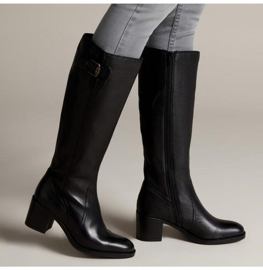 A pair of black leather knee-high boots with block heels, shown on a model wearing light gray jeans against a neutral beige studio background.