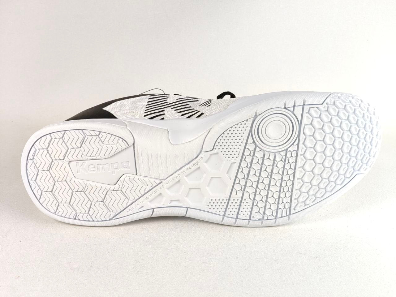 White Kempa indoor court shoe shown from the sole view, featuring a textured white outsole with hexagonal and chevron patterns, with a hint of black and white upper design above.