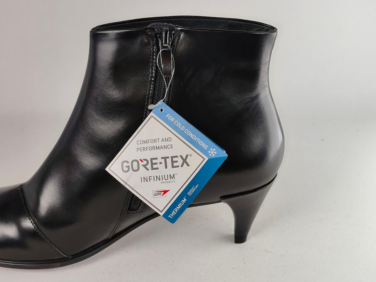 A black leather ankle boot with a pointed toe and a low kitten heel, featuring a side zipper and a hanging GORE-TEX Infinium tag, photographed against a neutral light gray background.