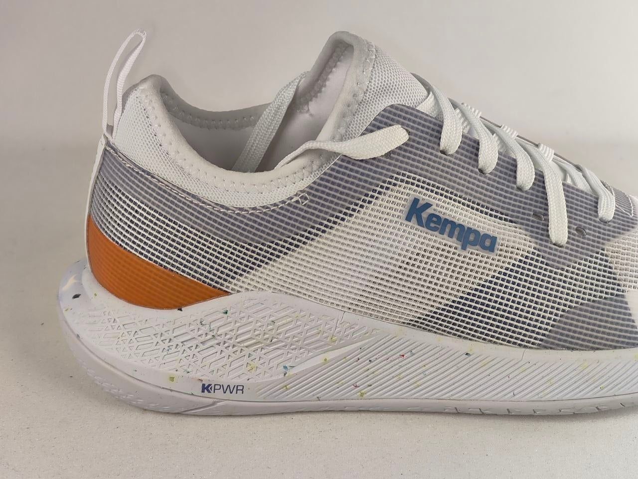 A white and gray mesh athletic sneaker with a textured orange heel accent and a blue 'Kempa' logo on the side, shown against a plain light background.