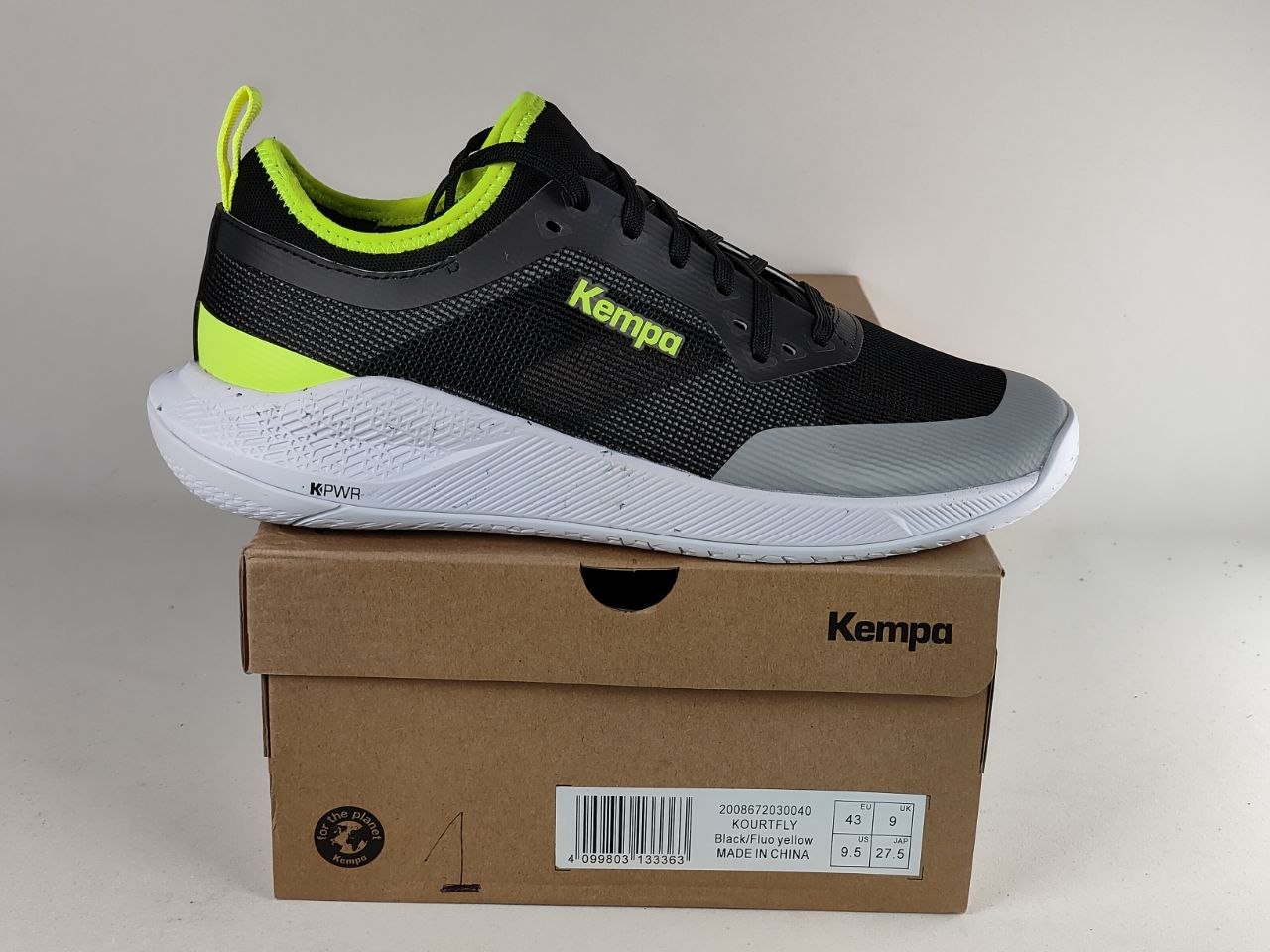A black and gray Kempa athletic sneaker with bright neon yellow-green accents, resting on top of its brown cardboard shoebox against a plain light background.