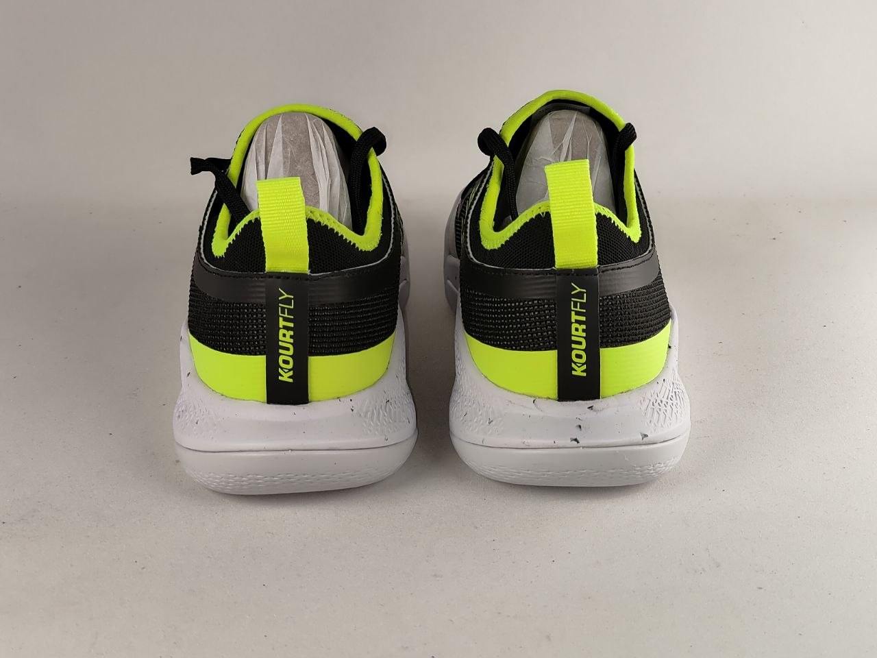 A pair of Kourtly athletic sneakers shown from the back, featuring a black and neon yellow-green color scheme with a white sole, set against a plain light gray background.