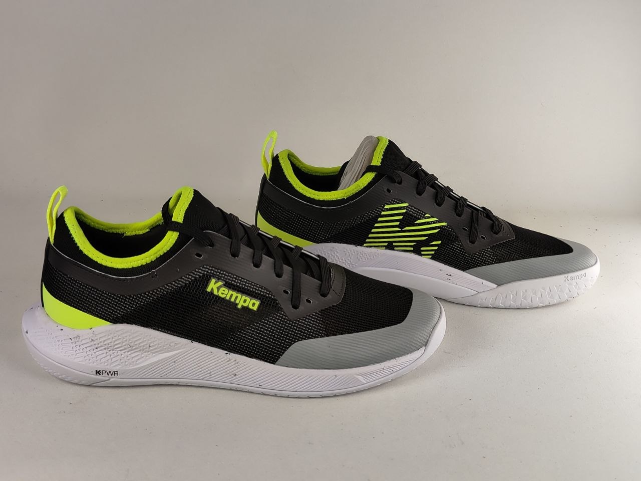 A pair of black Kempa athletic sneakers with bright neon yellow-green accents, gray toe caps, and white soles, displayed on a light gray studio background.