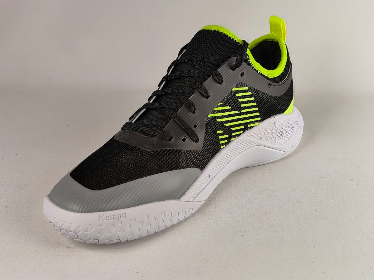 A single athletic sneaker shown from a three-quarters angle on a neutral light gray background. The shoe features a black mesh upper with gray overlays, a white textured midsole, and vibrant neon yellow-green accents on the side logo, collar lining, and heel pull tab.