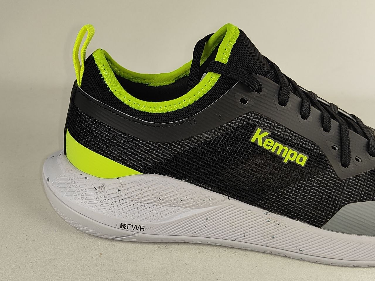 A close-up side view of a black and gray running shoe with bright neon yellow-green accents, featuring the Kempa logo on the side and a white textured midsole.