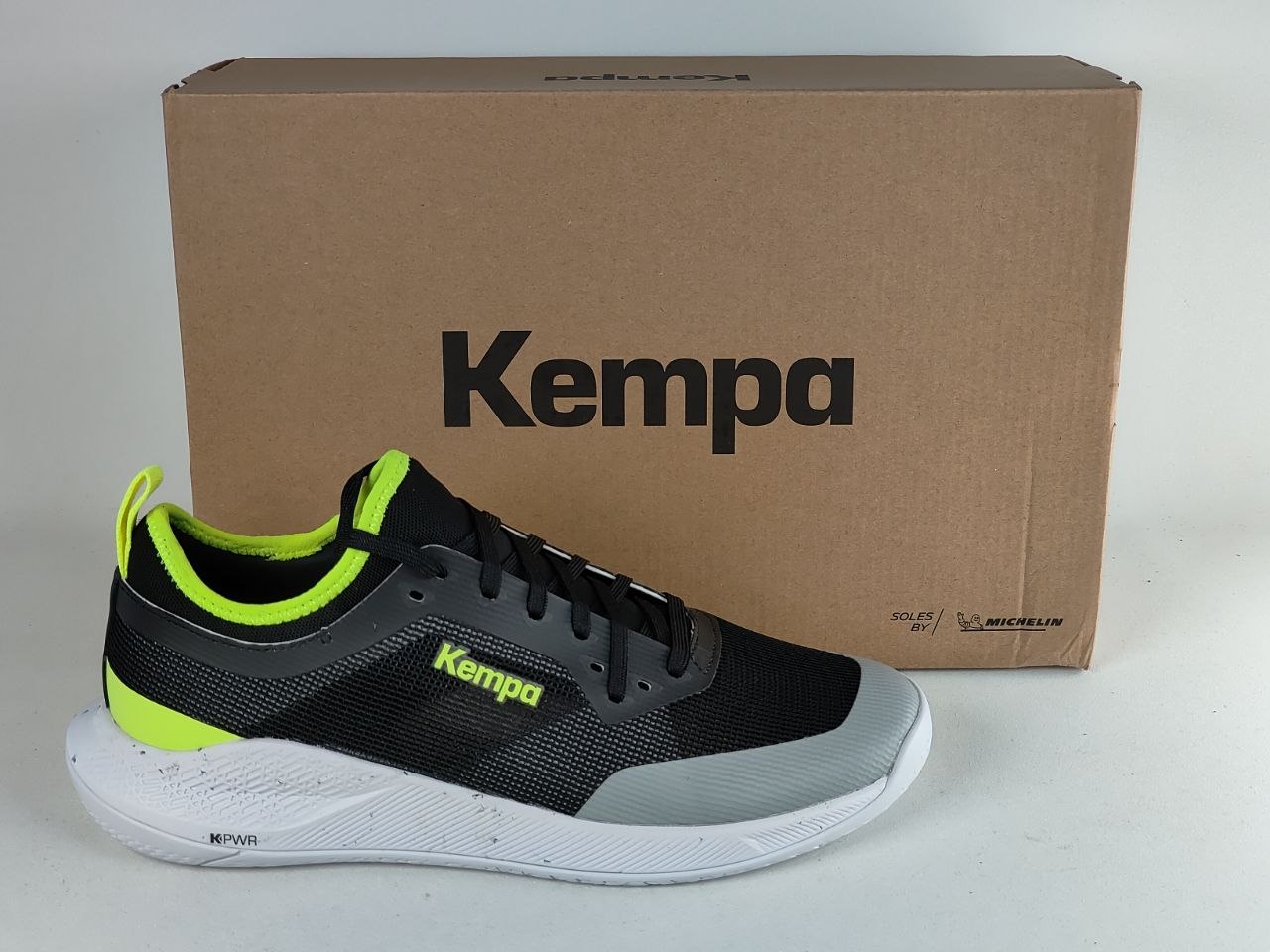 A black and gray Kempa athletic shoe with bright neon green accents, shown in front of its brown cardboard box on a light gray background.