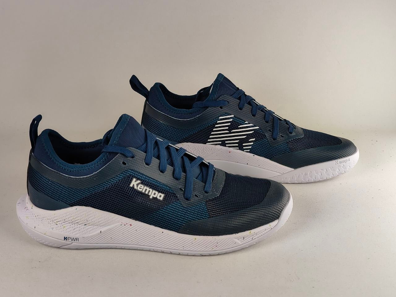 A pair of navy blue and white Kempa athletic sneakers, with one shoe positioned in front of the other on a plain light beige background.