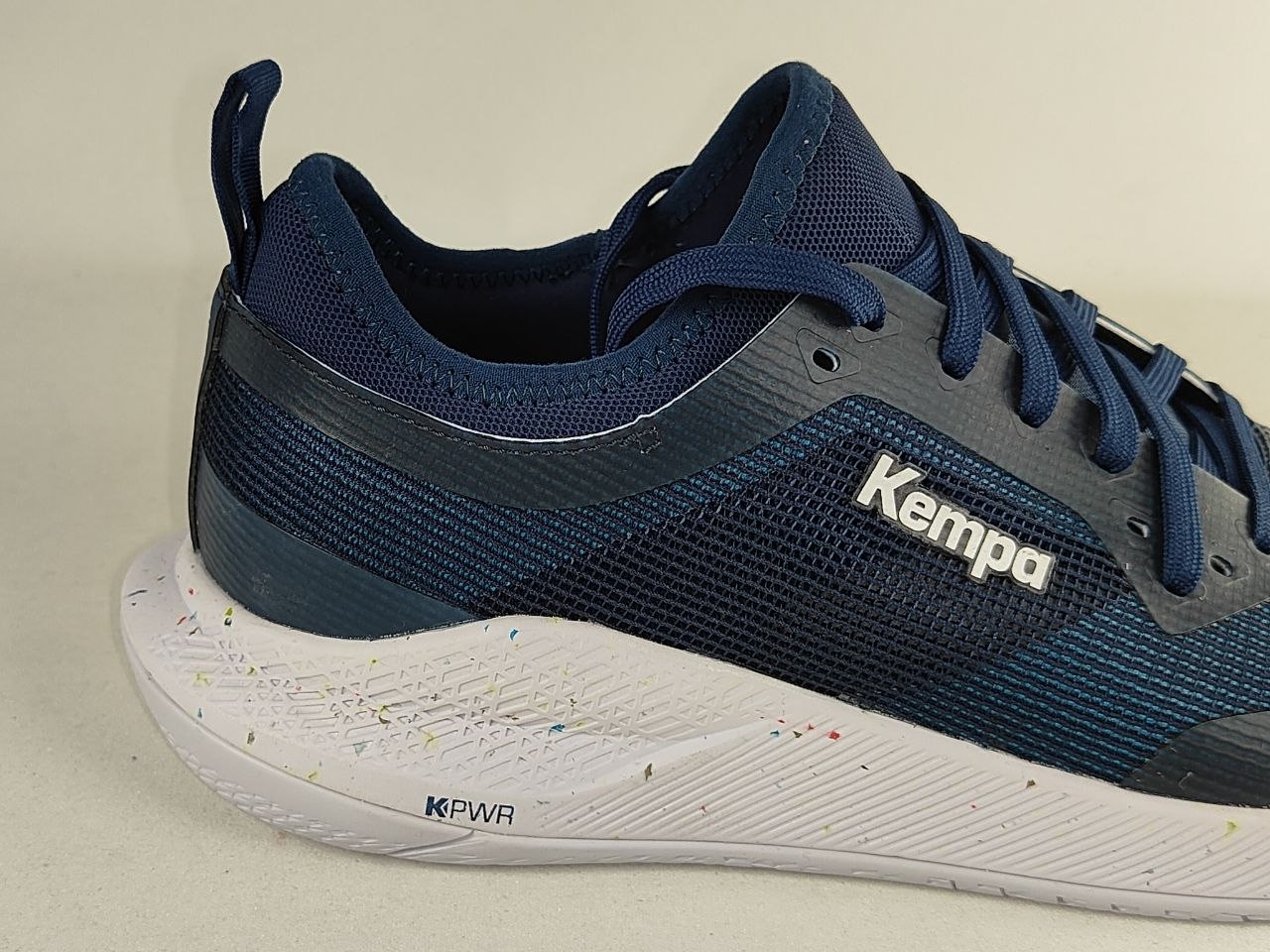 A close-up side view of a Kempa athletic shoe in navy blue and dark gray with a white speckled sole, on a light beige background.