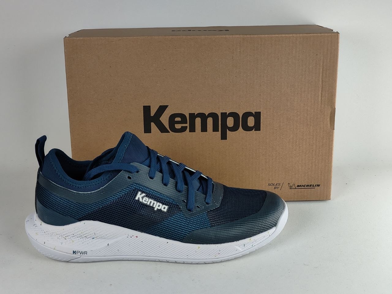 A Kempa athletic shoe in dark blue and navy with a white sole, positioned in front of its brown cardboard packaging box on a light gray background.