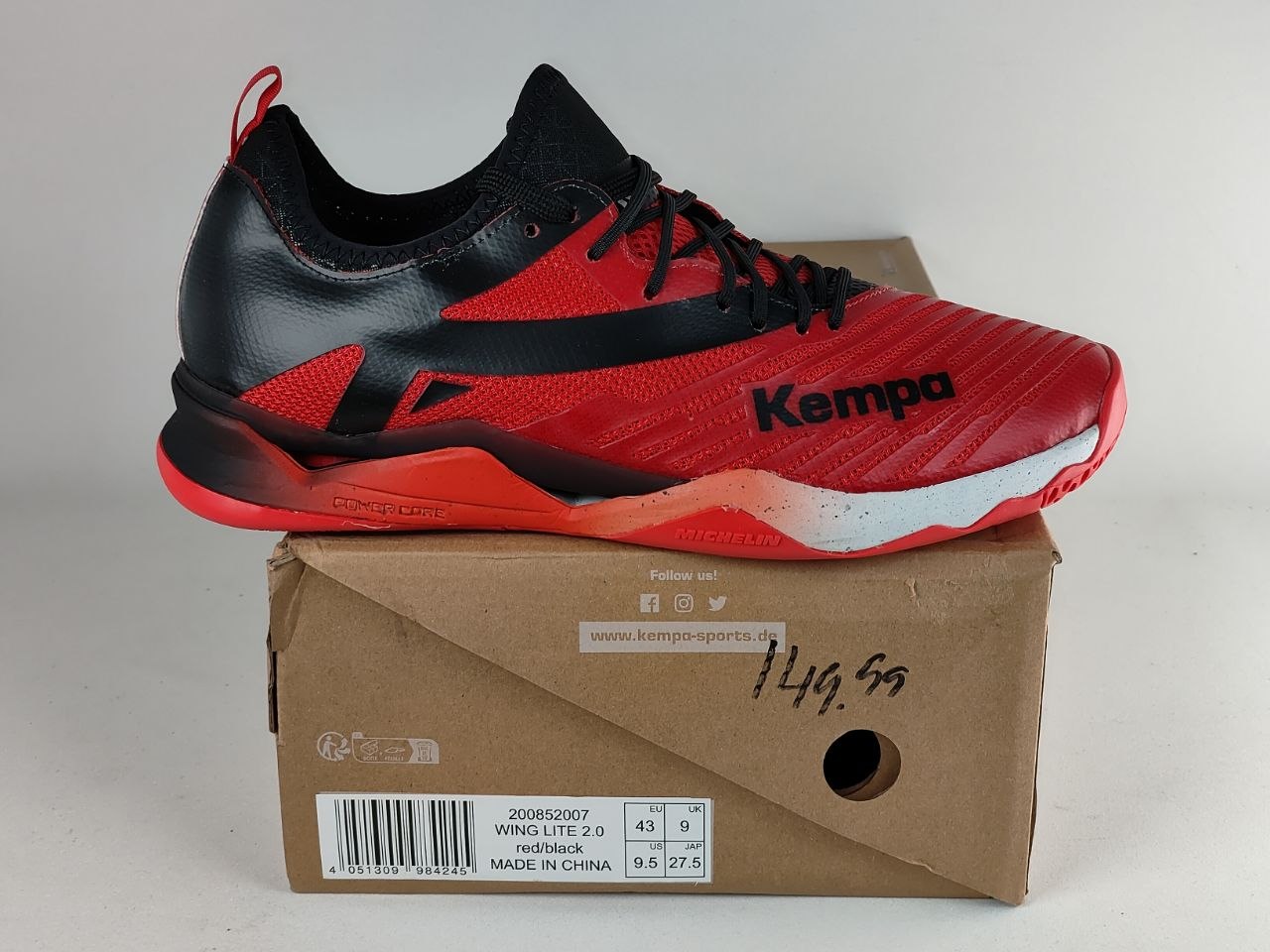 A red and black Kempa badminton shoe resting on its original brown cardboard box against a light gray background.
