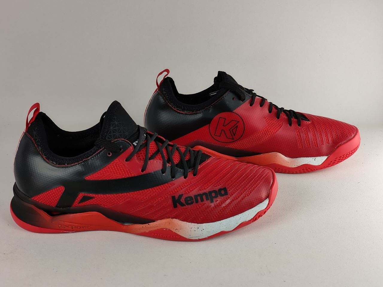 A pair of red and black Kempa athletic shoes with a textured synthetic upper, black laces, and a red and black sole, displayed on a light gray background.