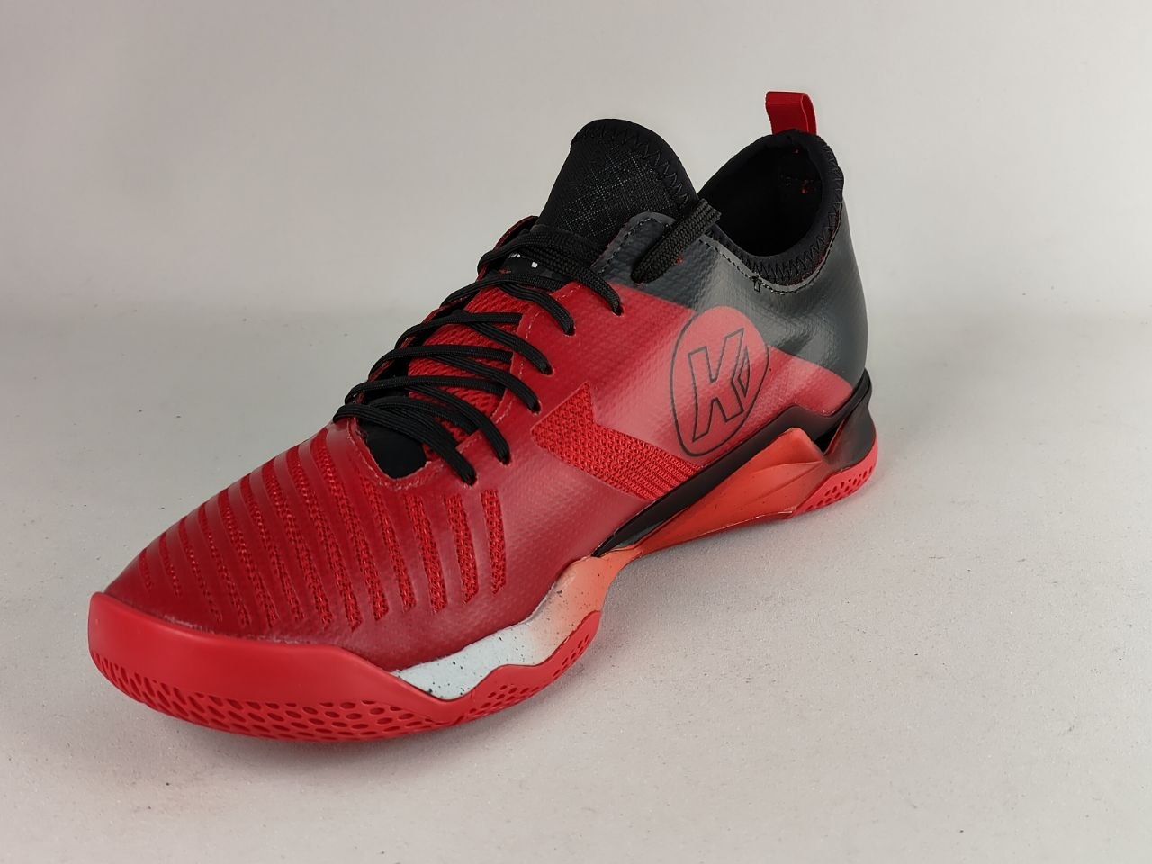 A red and black athletic shoe with a textured knit upper, black laces, and a red and black sole, shown on a neutral light gray background.