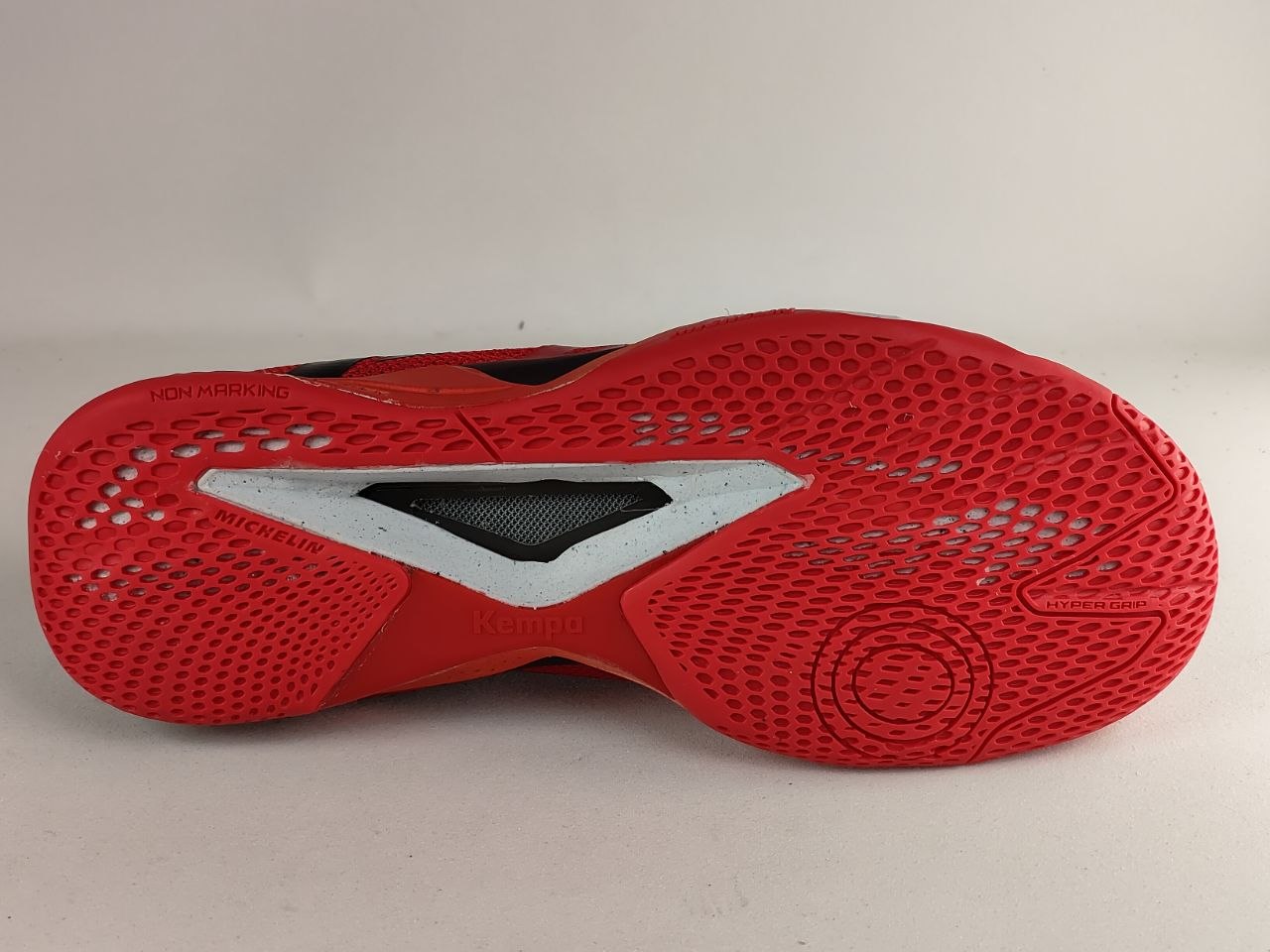 The outsole of a red and black athletic shoe, showing a textured red rubber tread with a white and black V-shaped accent in the center, photographed against a neutral light gray background.