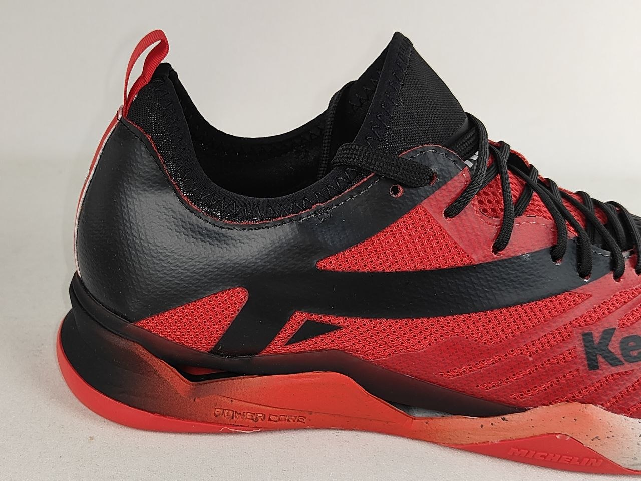 A close-up of a red and black athletic shoe, showing the side profile with black laces, a black collar, and a red and black sole on a light gray background.