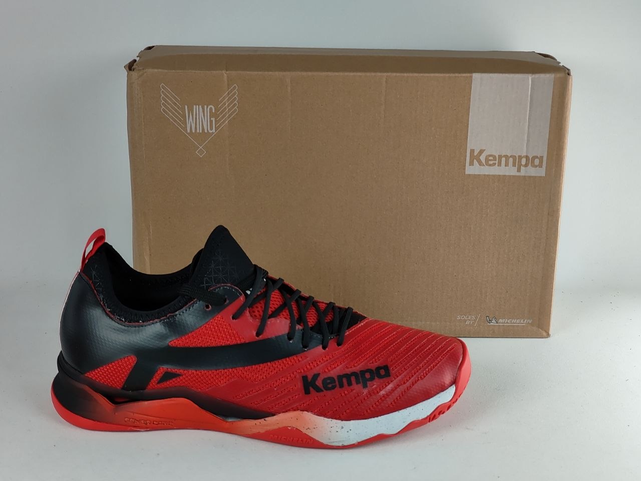 A red and black Kempa athletic shoe positioned in front of its brown cardboard packaging box on a light gray background.