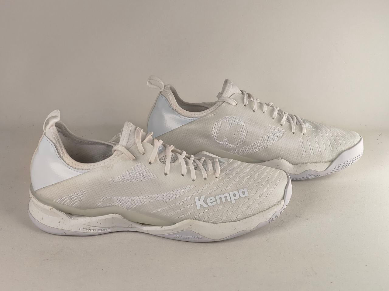 A pair of white Kempa athletic sneakers with a textured mesh upper and white laces, shown on a neutral light beige background.