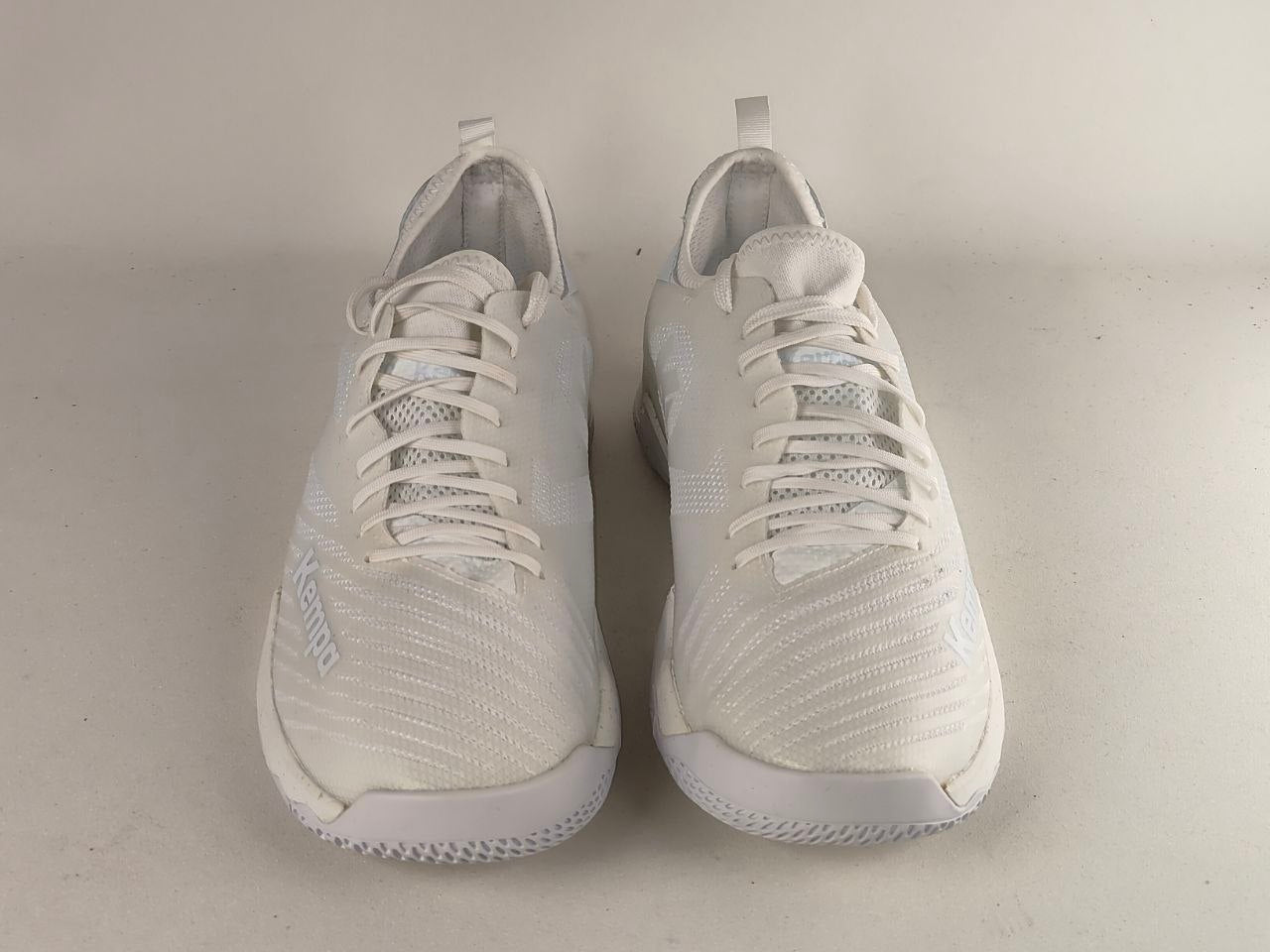 A pair of white athletic sneakers with textured knit uppers, white laces, and white soles, photographed against a plain light beige background.
