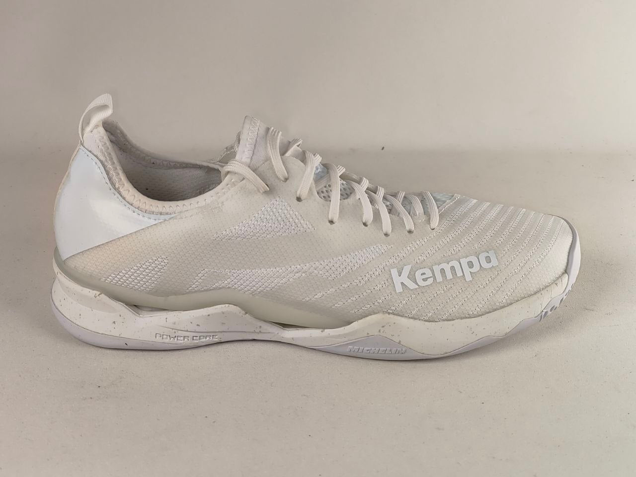 A white Kempa athletic shoe with a textured mesh upper, white laces, and a white sole, photographed against a neutral light beige background.