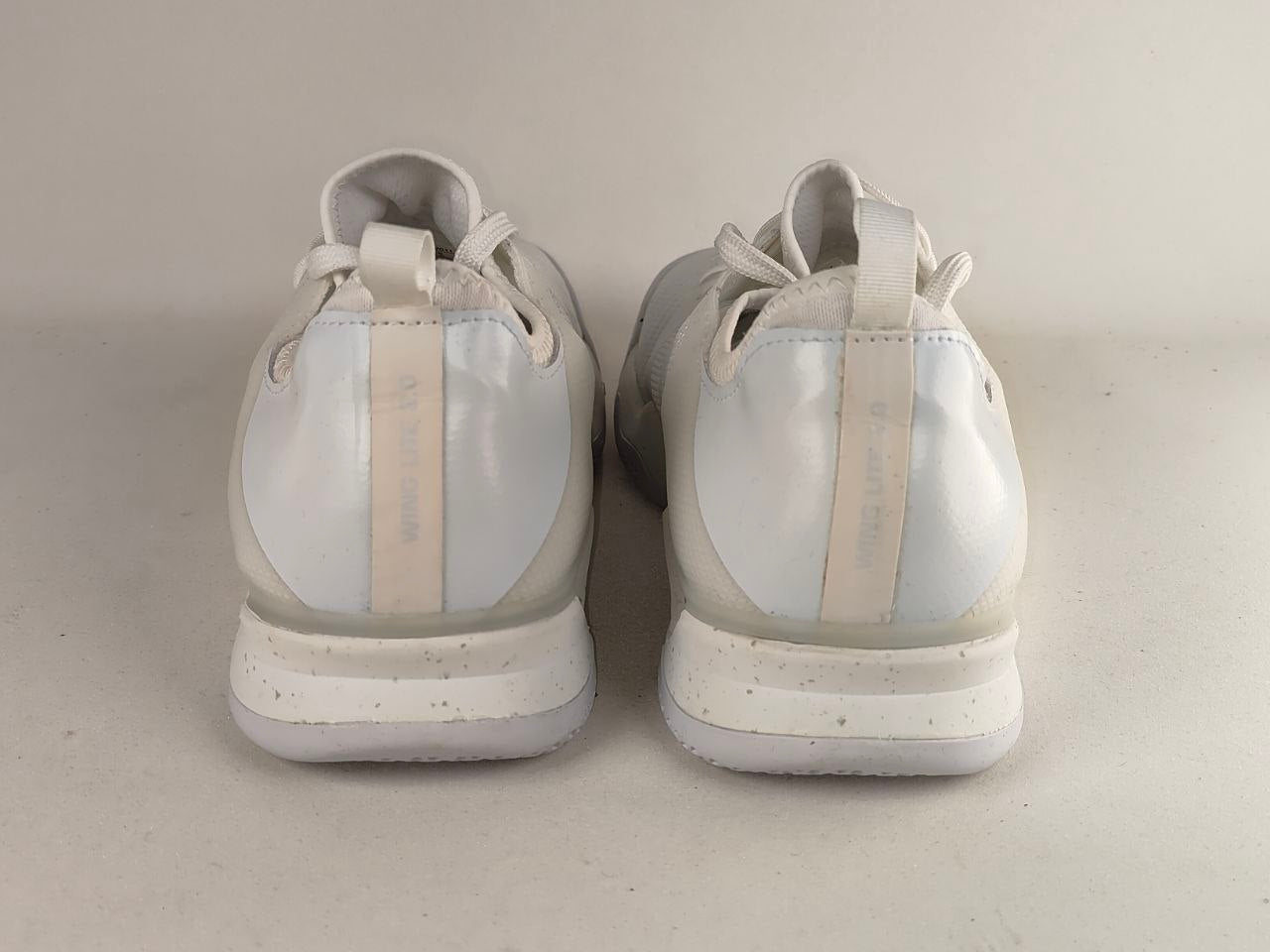 A pair of white low-top sneakers shown from the back, featuring a glossy white heel counter with a vertical beige stripe and white laces, set against a plain light gray background.