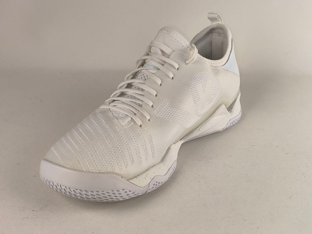 A white athletic sneaker with a textured knit upper, white laces, and a white sole, photographed on a neutral light gray background.