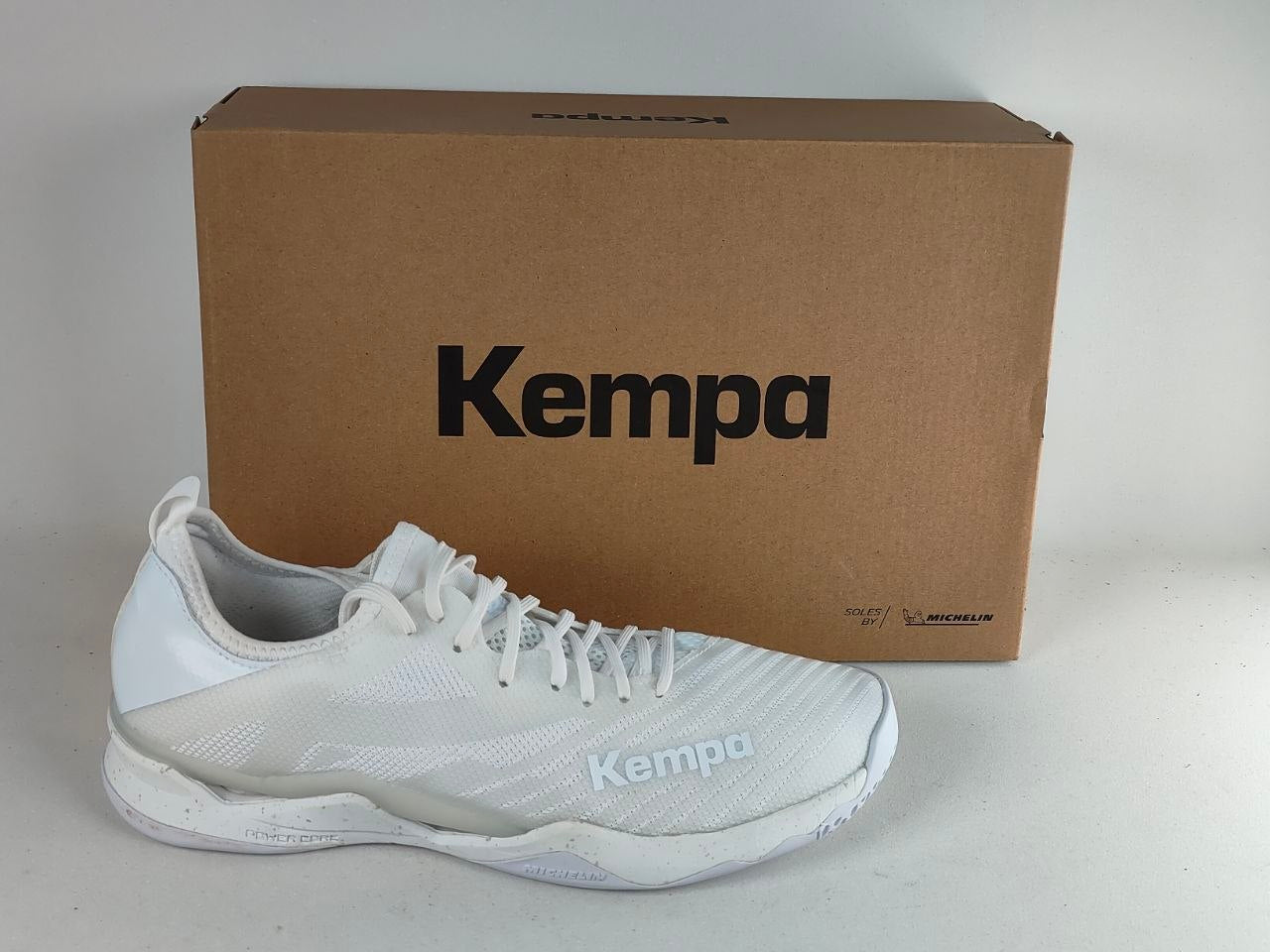 A white Kempa athletic shoe positioned in front of its brown cardboard packaging box, which features the black Kempa logo.