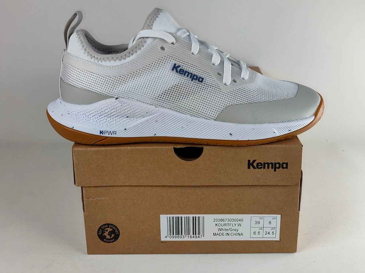 A white and light gray Kempa athletic shoe with a brown gum sole, displayed on top of its brown cardboard shoebox against a neutral light gray background.