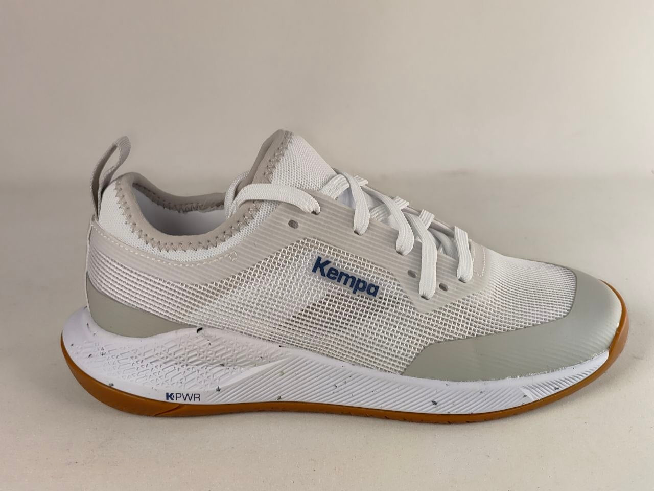 A white and light gray Kempa athletic shoe with a brown outsole, shown from a side profile view against a neutral beige background.