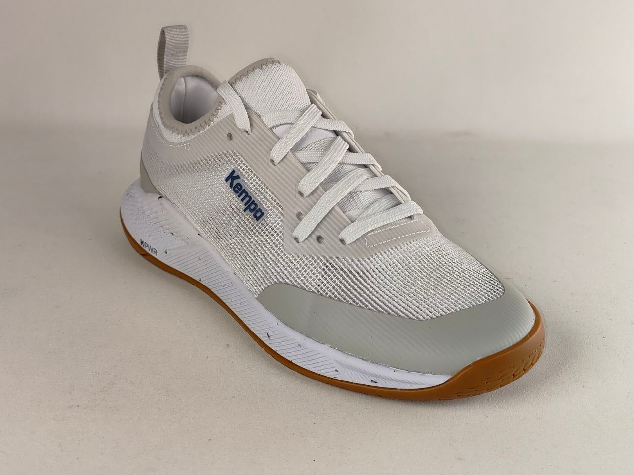 A white and light gray Kemper brand athletic shoe with white laces, a gray translucent toe bumper, and a brown outsole, photographed on a neutral beige background.
