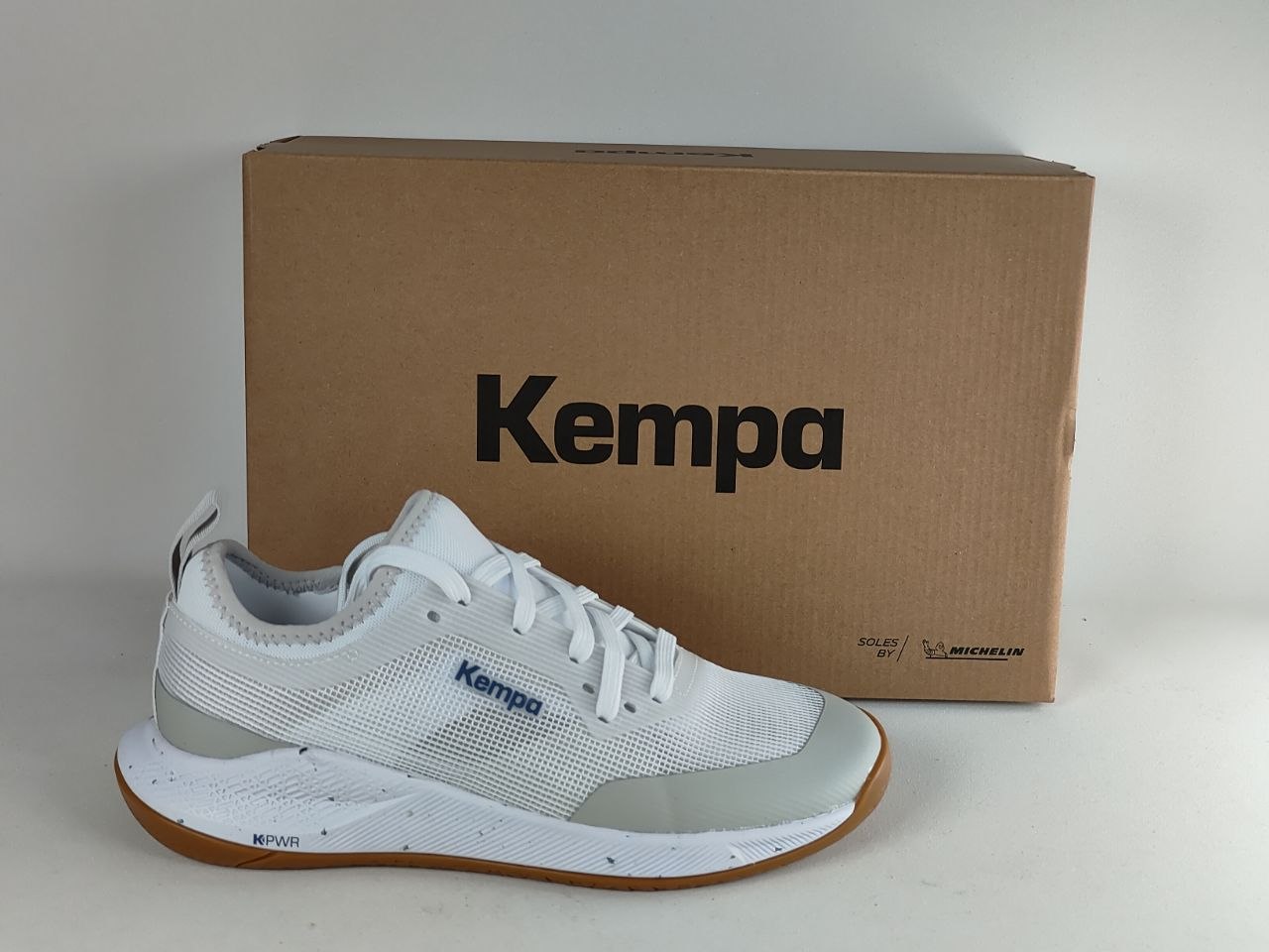 A white and light gray Kempa athletic shoe with a brown gum sole, positioned in front of its brown cardboard box on a light gray background.