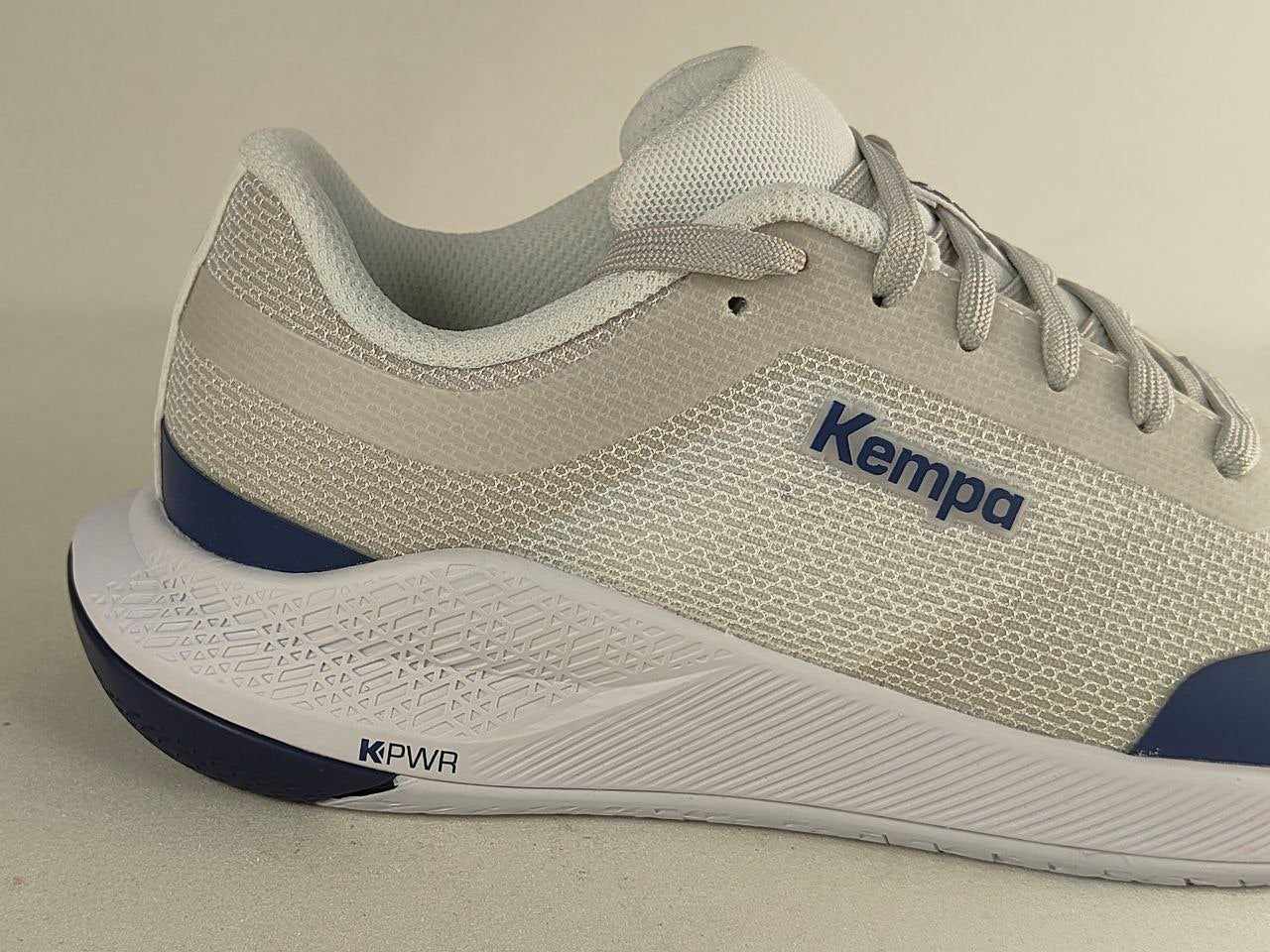 A close-up side view of a Kempa running shoe in light gray and white with navy blue accents on the midsole and toe cap.