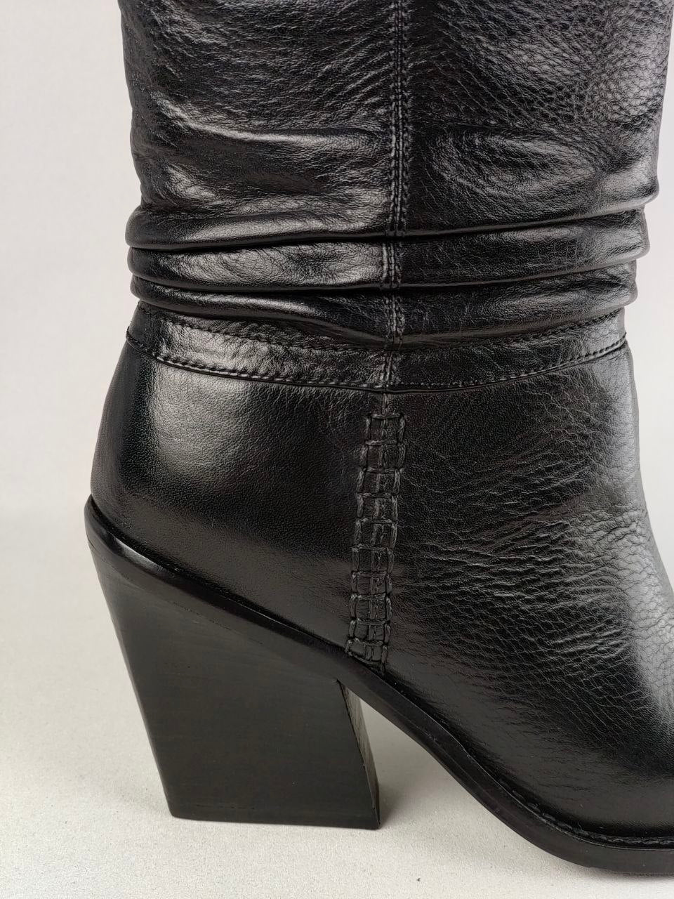 A close-up of a black leather boot with a textured grain finish, a ruched shaft, and a chunky block heel, shown against a light gray background.