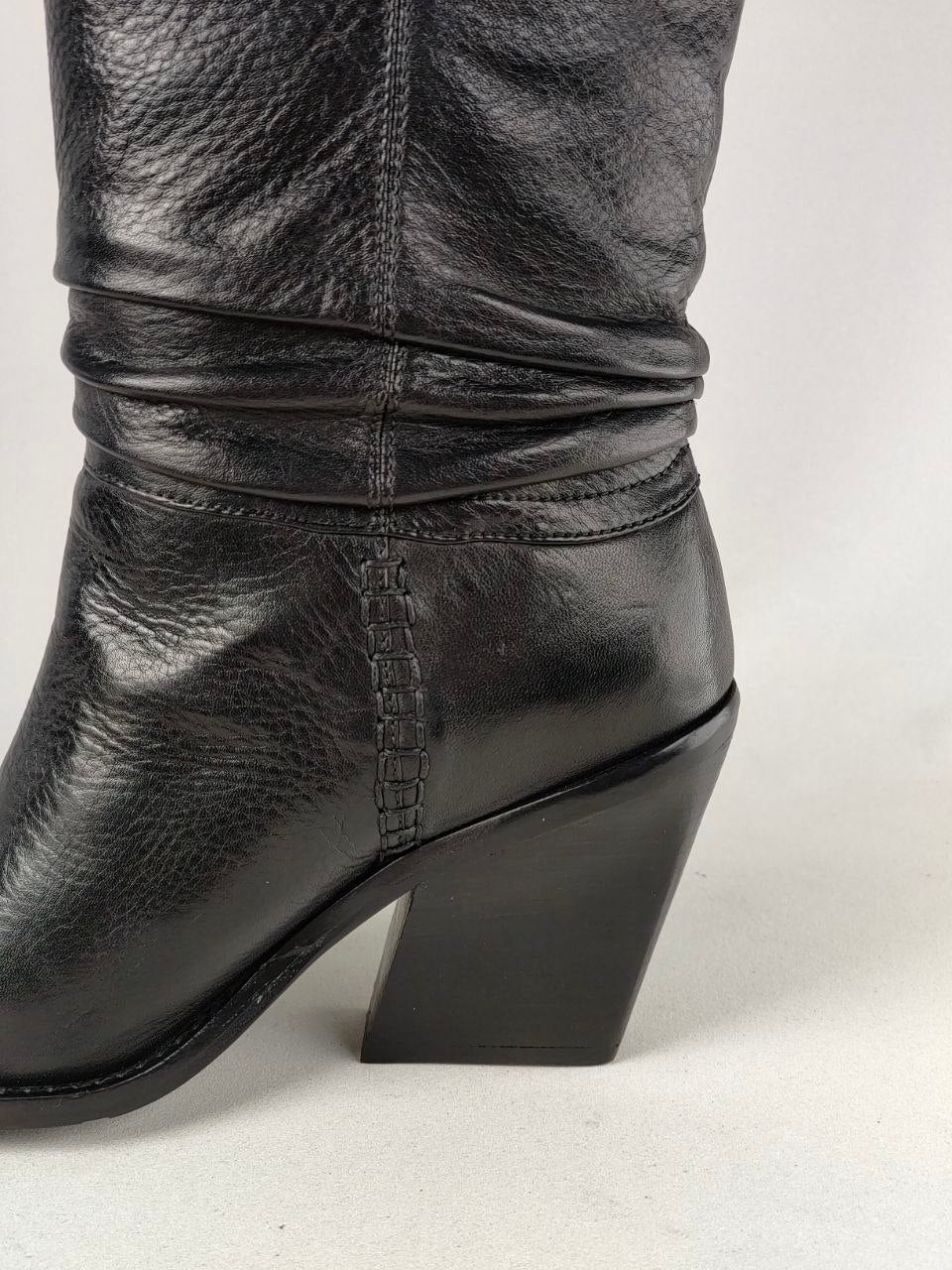 A close-up shot of a black leather boot with a chunky block heel, showing the side profile and textured leather grain against a light gray background.