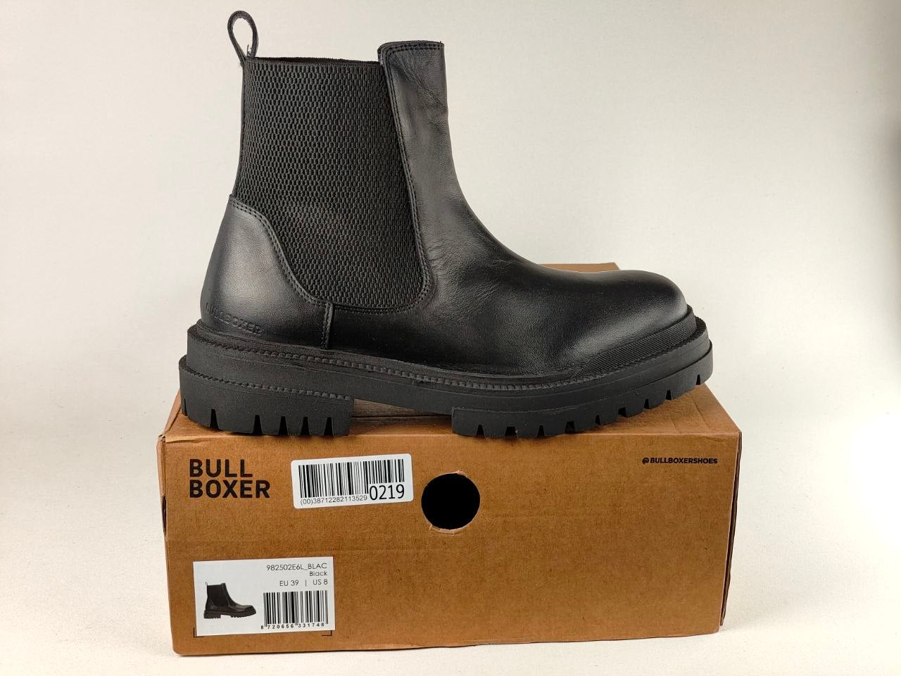 A black Chelsea-style ankle boot with a thick lug sole, shown standing on top of a brown Bull Boxer shoebox against a plain light background.
