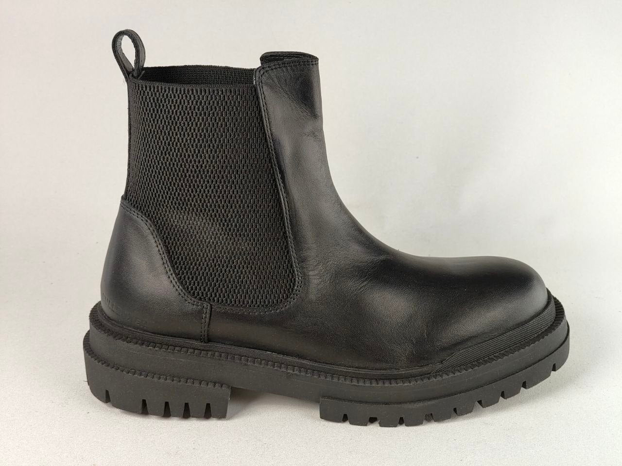 A black leather Chelsea-style ankle boot with a black elastic side panel and a chunky lug sole, shown from a side profile against a light gray background.