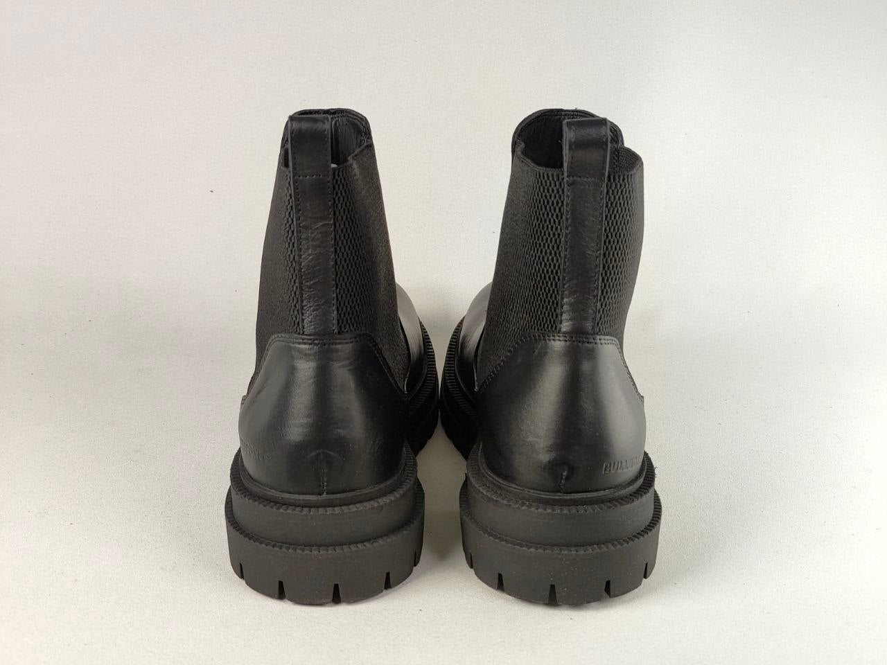 A pair of black Chelsea boots shown from the back, featuring smooth leather panels, textured elastic side panels, and chunky lug soles, set against a plain light background.