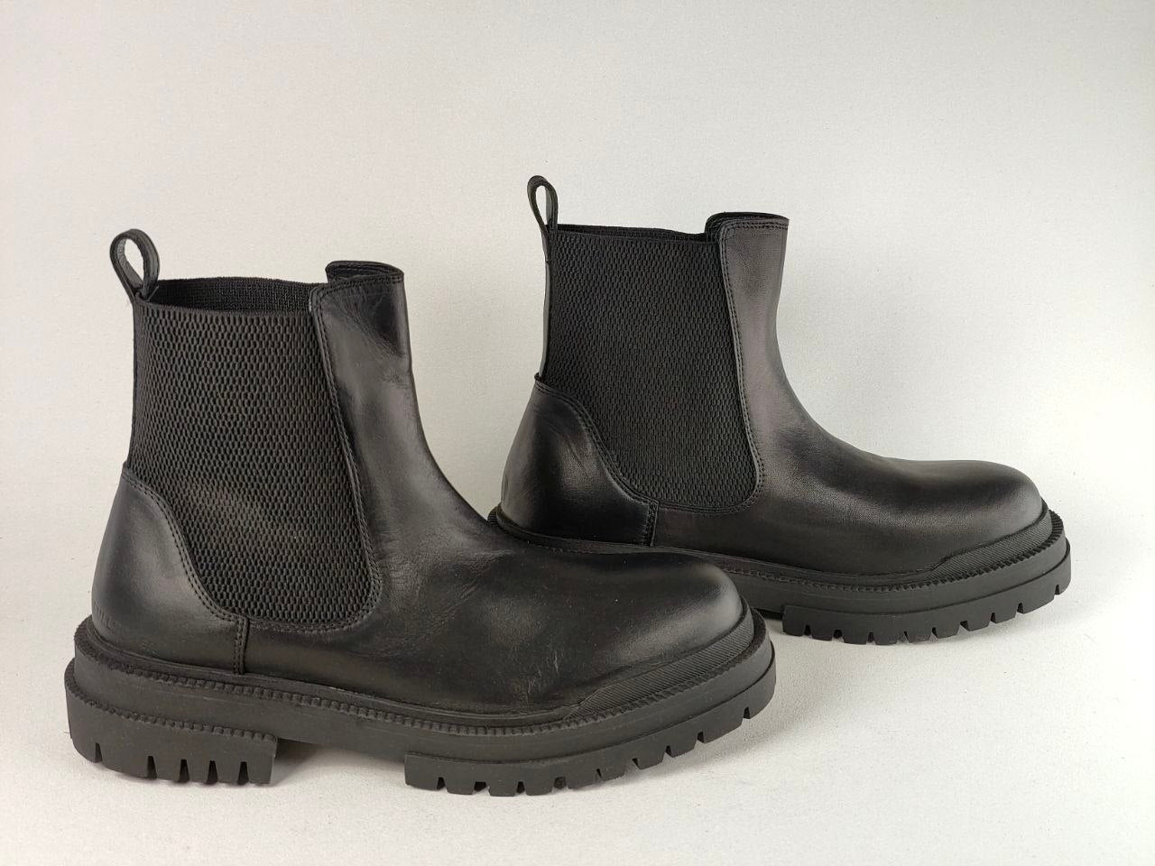 A pair of black leather Chelsea boots with black elastic side panels and chunky lug soles, photographed on a light gray studio background.
