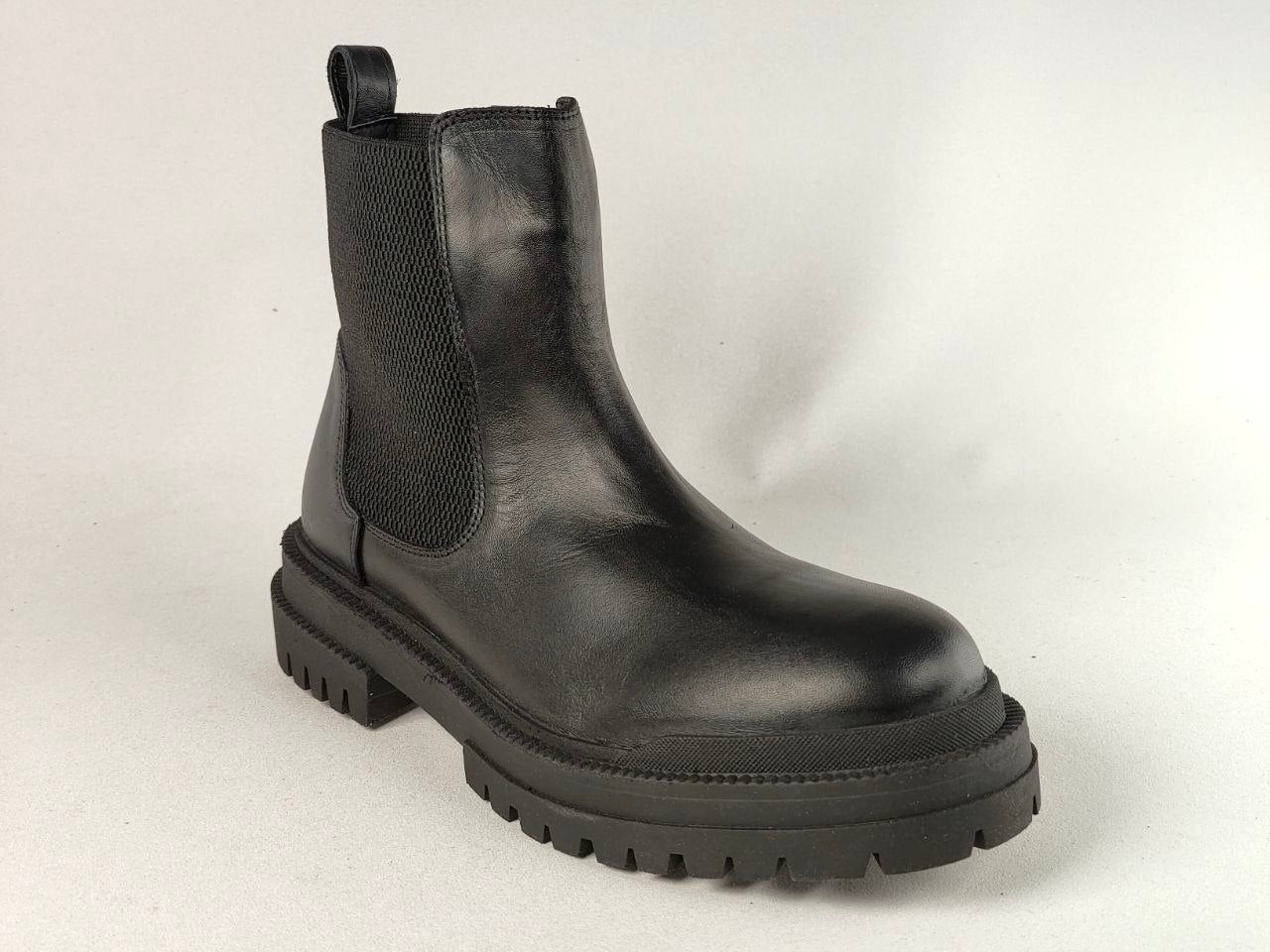 A black leather Chelsea boot with a chunky lug sole and elastic side gore, photographed on a light gray studio background.