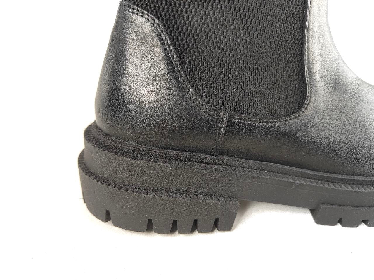 Close-up of the side and heel of a black leather Chelsea boot with a black elastic side panel and a chunky lug sole, shown on a white background.