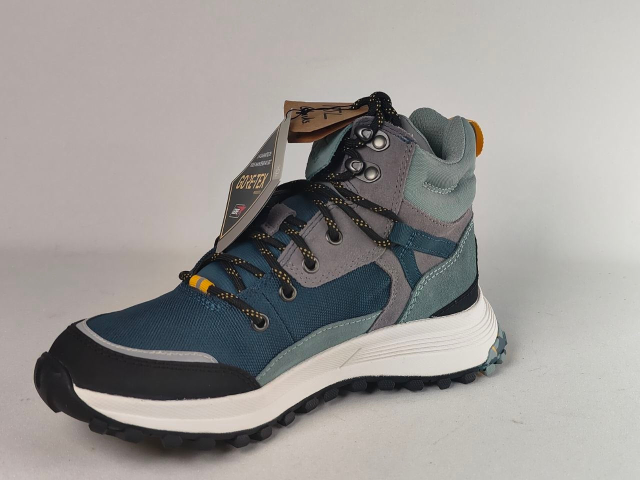 A mid-cut hiking boot in slate blue, gray, and black with a white midsole, shown with its paper tag attached against a neutral gray background.