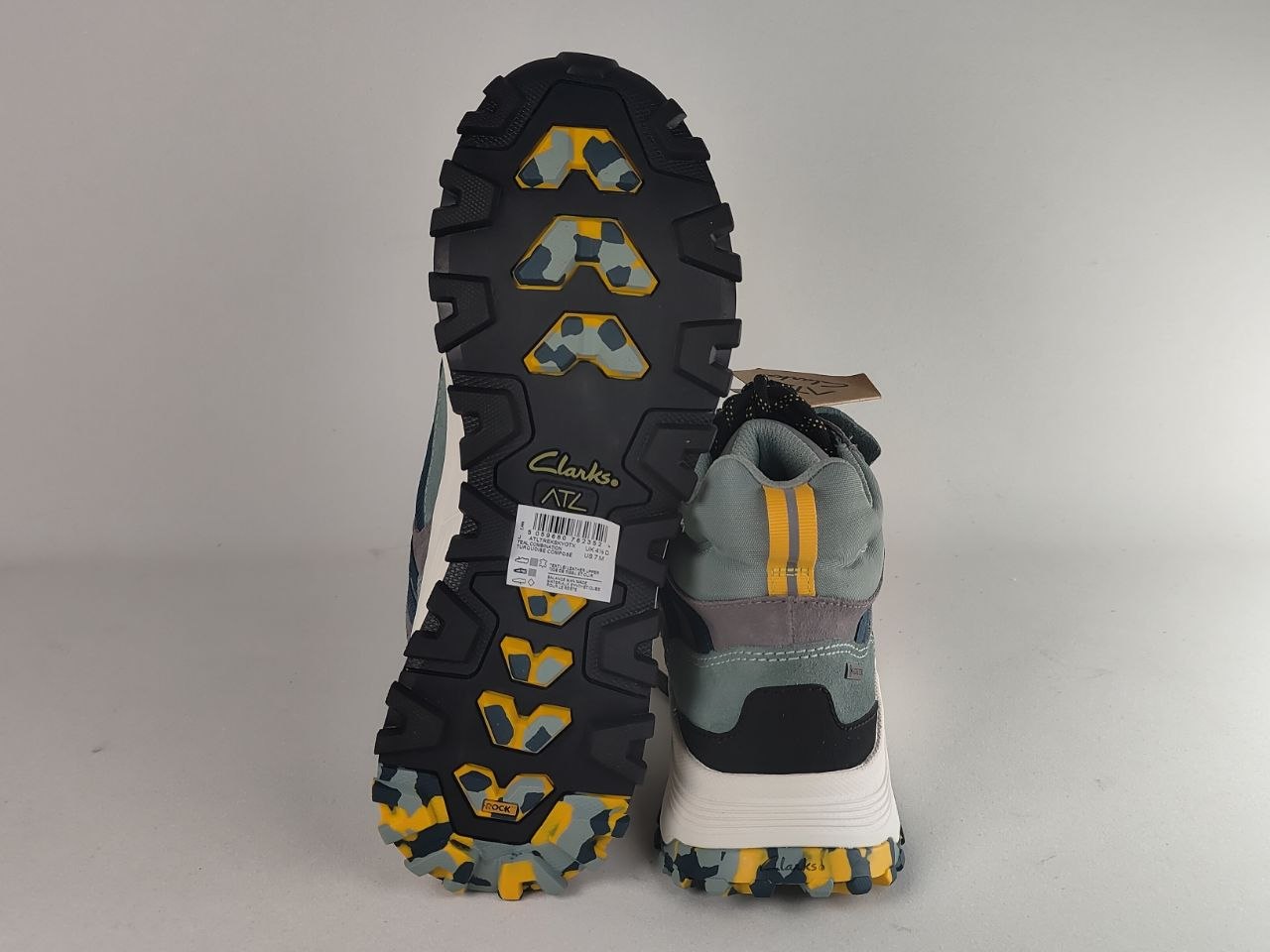 A pair of Clarks N72 hiking shoes shown against a neutral light gray background, with one shoe standing upright to display the black treaded sole and the other lying down to show the side profile. The shoes feature a color-blocked design in sage green, gray, black, white, and yellow accents.