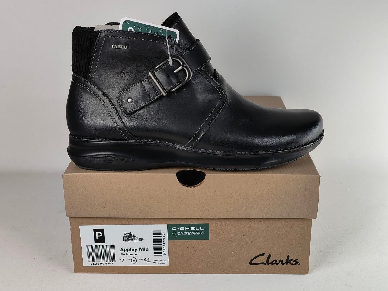 A black leather ankle boot with a buckle strap, displayed on top of its brown Clarks shoebox against a light gray background.