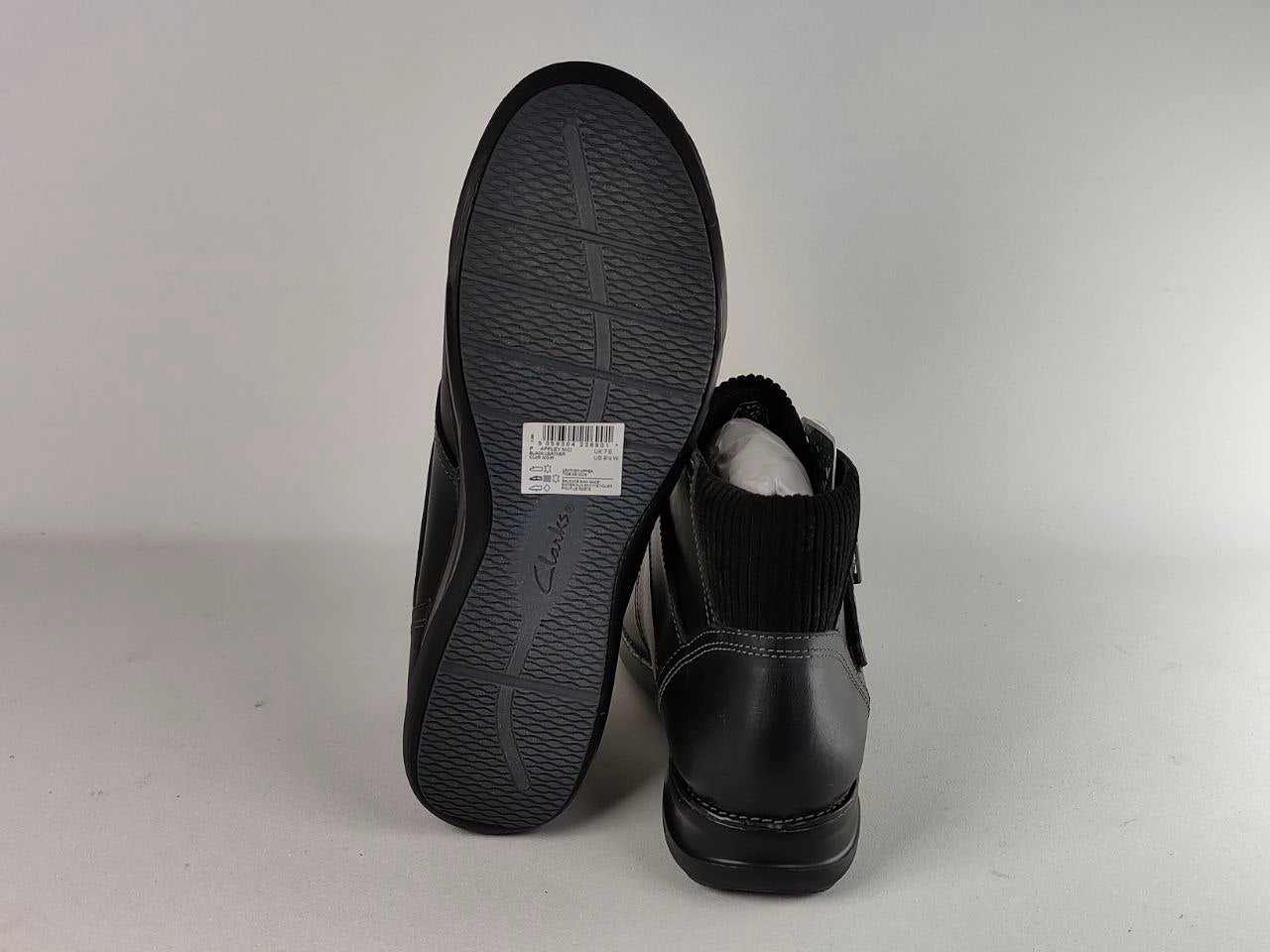 A pair of black ankle boots shown on a light gray background, with one boot standing upright to display the black textured sole and the other lying on its side showing the black knit collar and black leather upper.