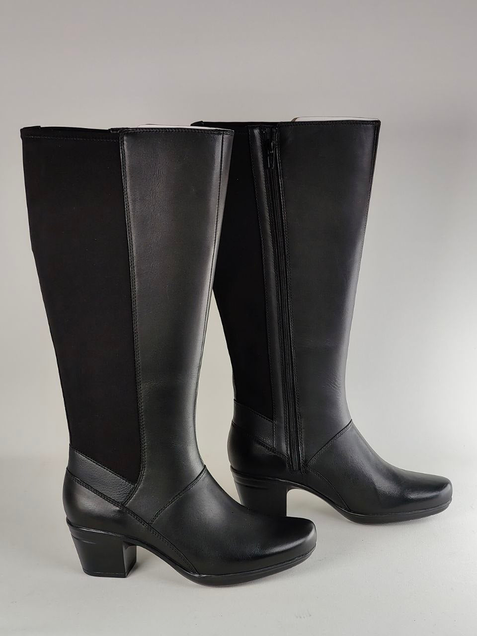 A pair of black knee-high leather boots with side zippers, shown against a neutral gray studio background.