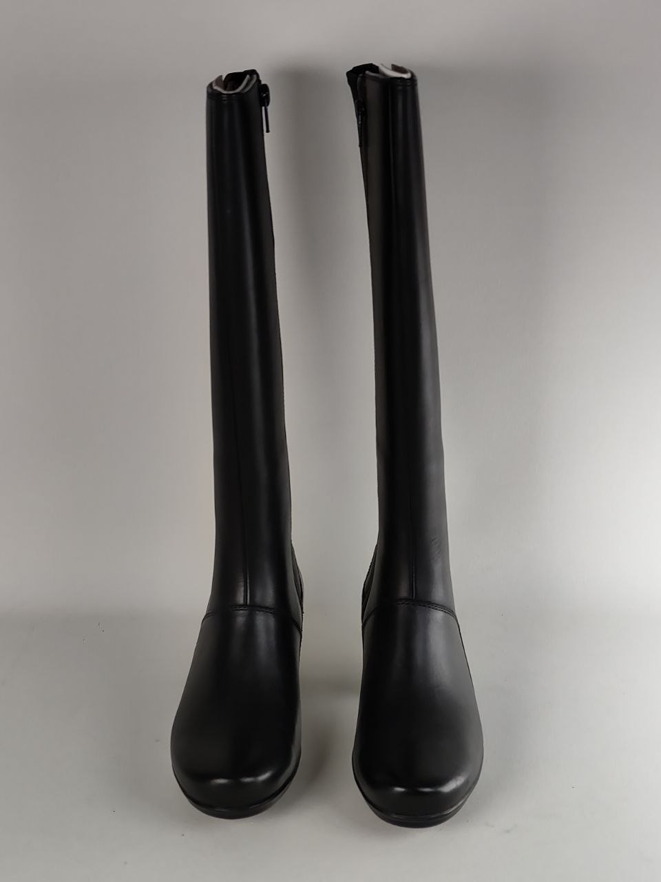 A pair of black knee-high leather boots with side zippers, shown from the front against a neutral gray background.